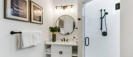 A bathroom with a white vanity, a round mirror, a towel rack, and a shower with a handheld showerhead.