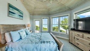 A bedroom with a large bed, a TV, wooden ceiling, and a view of the ocean.