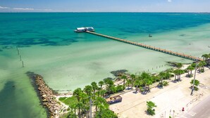 A coastal area with a pier extending into turquoise waters, a beach with palm trees, and a small structure near the water's edge.
