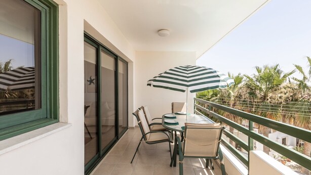 A balcony with a table and chairs, a striped umbrella, and a view of palm trees. - Mediterranean dream (Oroklini)