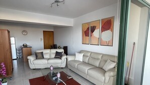 A modern living room with two white leather sofas, a glass coffee table, and a red rug. There is a dining area with a wooden table and chairs, and a kitchen area with cabinets and a countertop. - Mediterranean dream (Oroklini)
