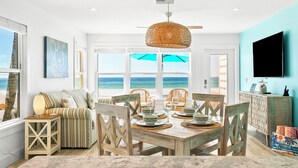 A dining area with a wooden table set for four, wicker chairs, and a view of the ocean through large windows.