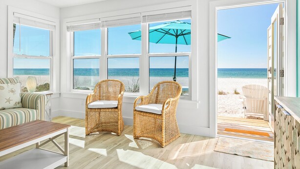 A room with large windows overlooking a beach, wicker chairs, a striped sofa, and a wooden table.