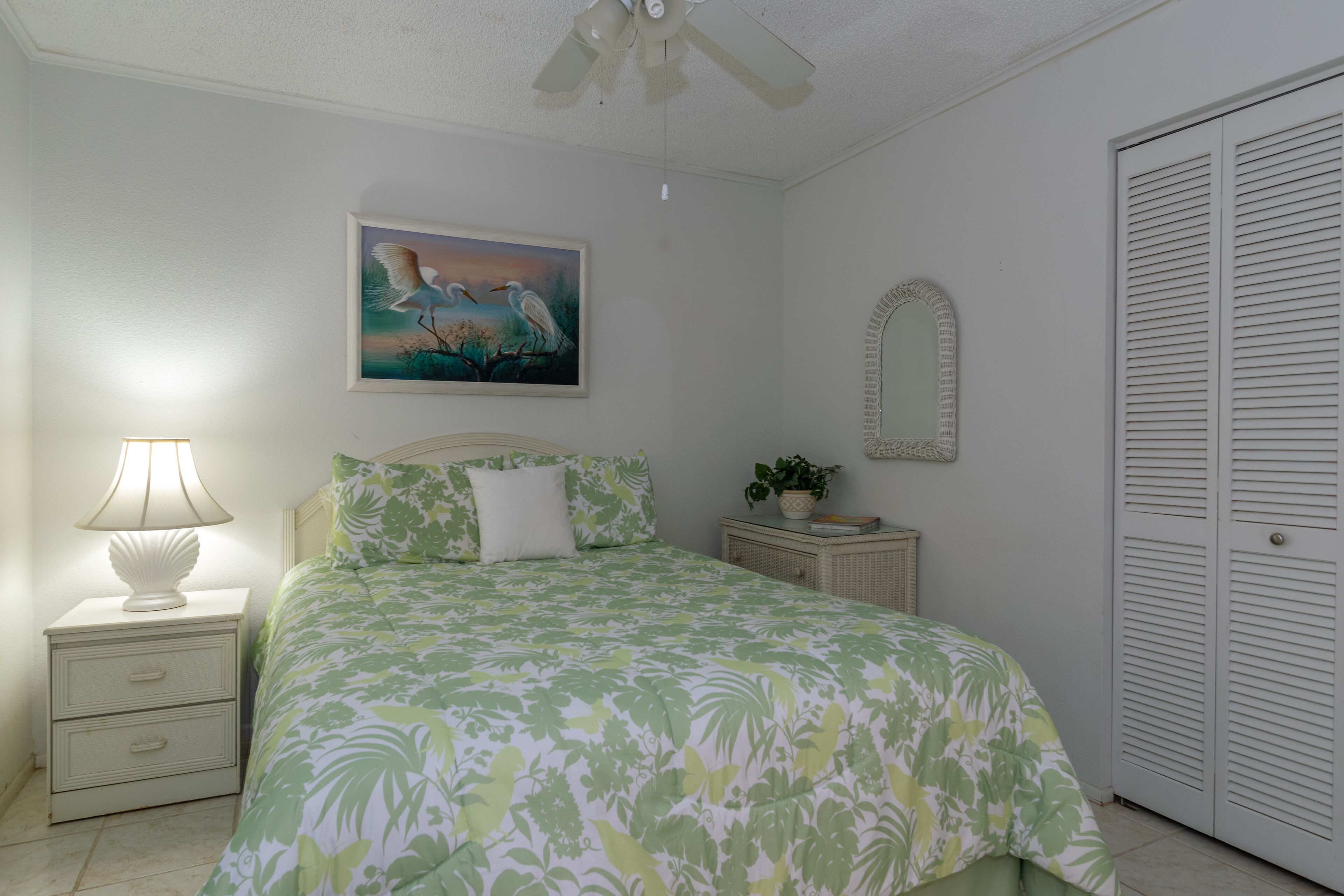 A bedroom with a bed, a nightstand with a lamp, a painting, a mirror, and a closet.