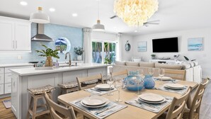 A modern kitchen with a central island, dining area with chairs and a table set for six, and a living area with a sofa and TV.