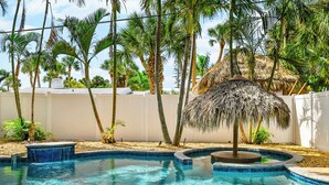 A swimming pool with a thatched umbrella, surrounded by palm trees and a white wall.