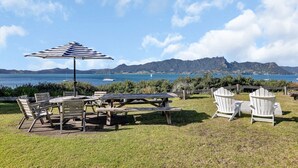An outdoor seating area with picnic tables and Adirondack chairs, overlooking a body of water and distant mountains.