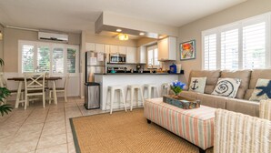 Una cocina moderna con armarios blancos, un refrigerador de acero inoxidable y un microondas. Hay un comedor con una mesa y sillas de madera, y una sala de estar con un sofá y reposapiés.