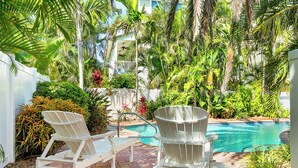 A poolside area with two Adirondack chairs, a white fence, and lush tropical plants. - Anna Cabana Bungalow #1. Steps to Gulf.  Shared Heated Pool. (Holmes Beach)