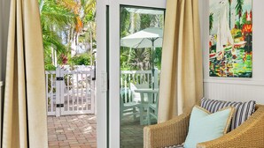 A room with a wicker sofa, a painting, and a view of a patio with chairs and umbrellas. - Anna Cabana Bungalow #1. Steps to Gulf.  Shared Heated Pool. (Holmes Beach)