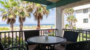A balcony with a round table and chairs, overlooking a beach with palm trees and a view of the ocean.