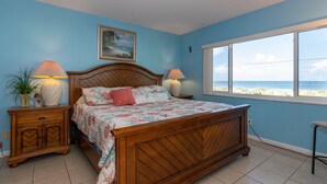 A bedroom with a wooden bed, two bedside tables with lamps, a nightstand, a window with a view of the beach, and a painting on the wall.