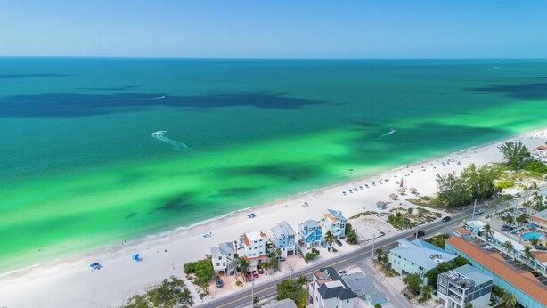 An aerial view of a coastal area with a beach, clear turquoise waters, and a row of buildings along the shore. - Bay Watch 6A · Dock · Bay & Gulf Access (Bradenton Beach)