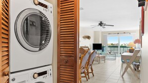 A laundry room with stacked washers and dryers, a dining area with chairs and a table, and a view of the outdoors through large windows. - Bay Watch 6A · Dock · Bay & Gulf Access (Bradenton Beach)