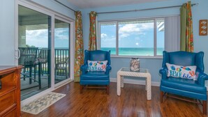 A living room with two blue armchairs, a small table, and a view of the ocean through large windows.