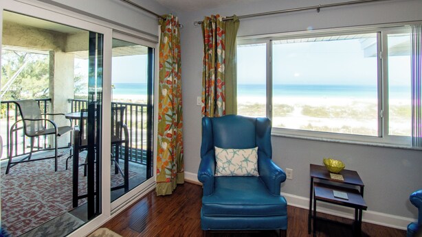 A room with a blue armchair, a small table, and a view of the beach through large windows.