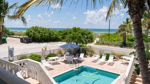 A pool area with a view of the beach, surrounded by palm trees and a white fence.