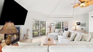 A spacious living room with a large white sofa, a wooden coffee table, and a ceiling fan. There are two windows with white blinds, a fireplace, and a TV mounted on the wall.