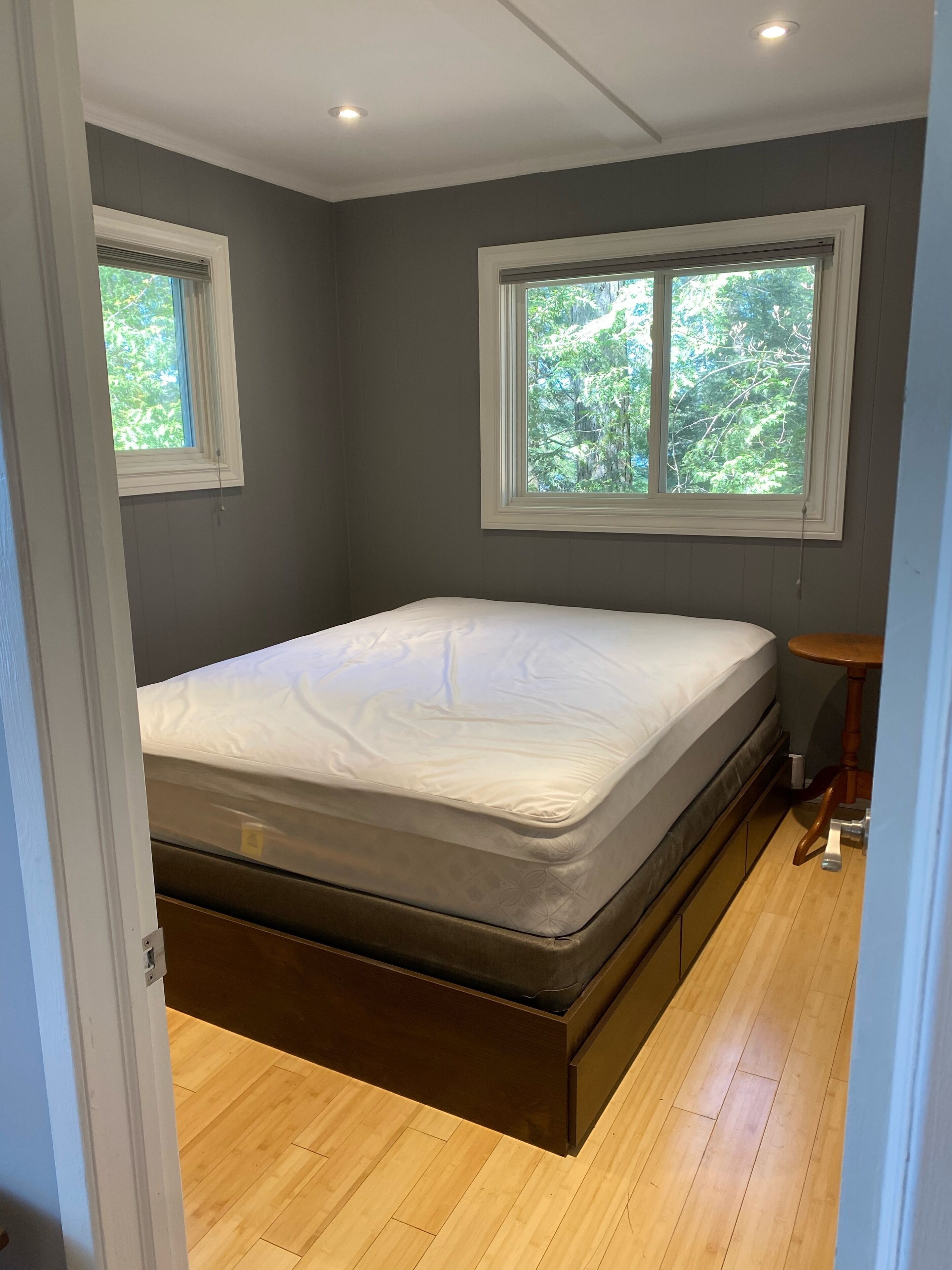 A bedroom with a bed, two windows, a small table, and a wooden floor.