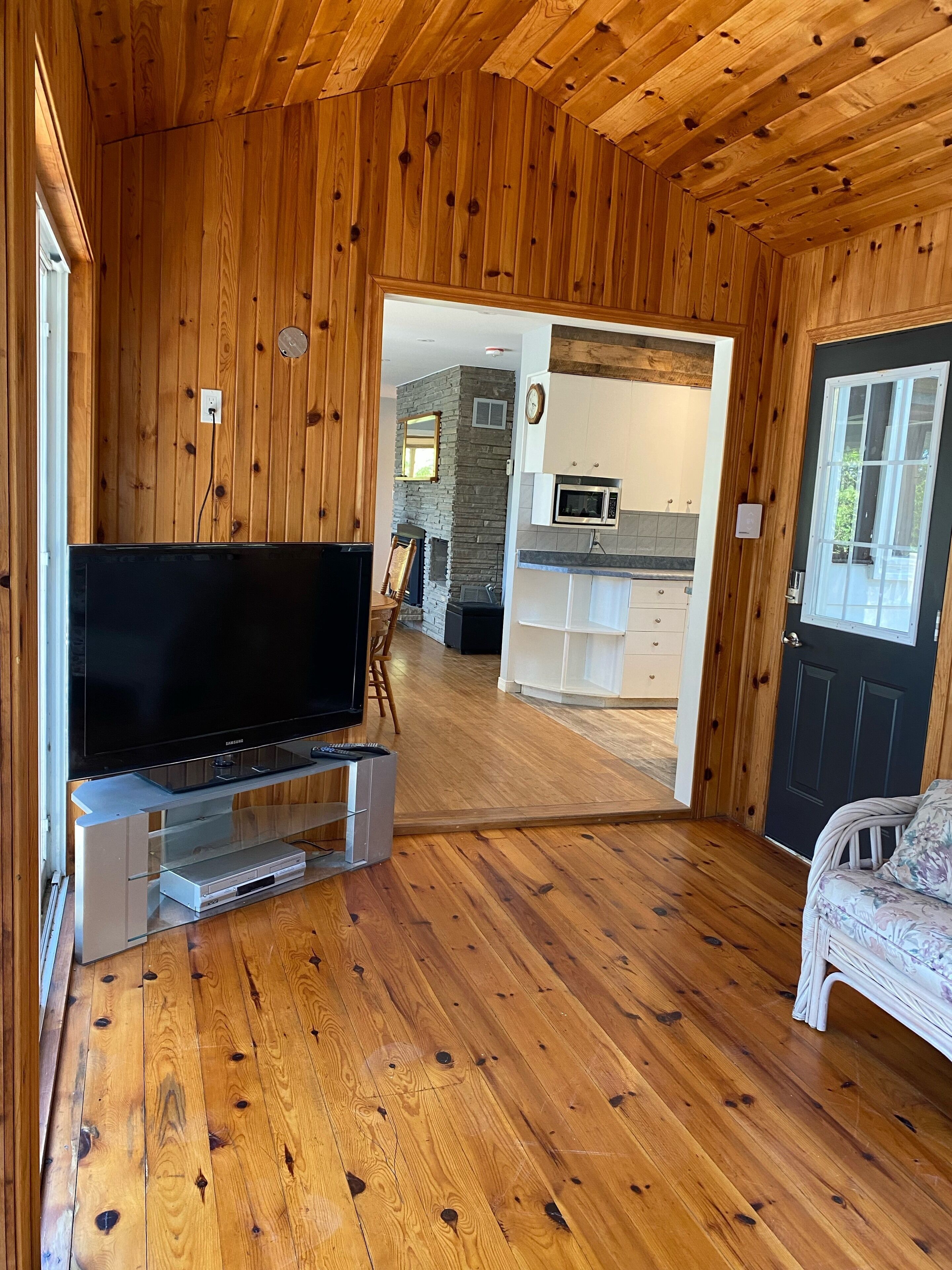 A wooden interior with a flat-screen TV on a stand, a stone fireplace, and a kitchen area with white cabinets and a microwave.