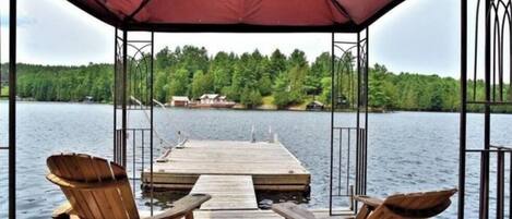 A wooden deck with two Adirondack chairs, a covered area, and a view of a lake and forest.