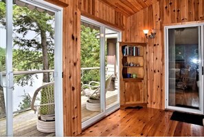 A wooden interior with a sliding glass door leading to a deck, a small bookshelf, and a cushioned chair. - Private 4 Season Waterfront Cottage - 2 hrs from TO -Great Swimming / FAST WI-FI (Minden)