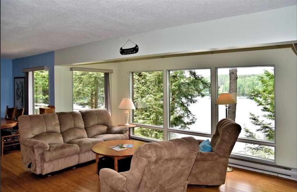 A living room with a sectional sofa, a recliner, and a coffee table. Large windows provide a view of trees and a body of water.