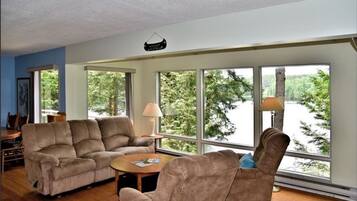 A living room with a sectional sofa, a recliner, and a coffee table. Large windows provide a view of trees and a body of water.