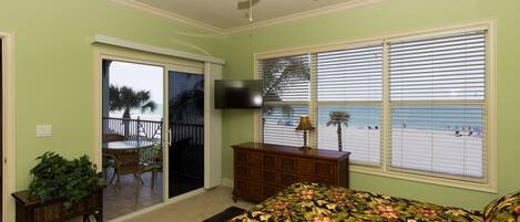 A bedroom with a bed, a ceiling fan, a television, a dresser, and a view of the beach through the window.