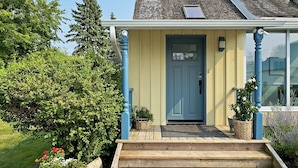 Una casa con puerta azul, un porche delantero de madera y un pequeño jardín con plantas en macetas.
