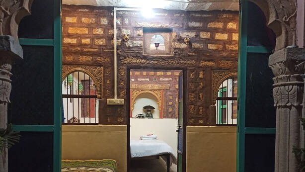 Ein Zimmer mit einem Bett, einem kleinen Tisch und einer Bank. Eine Tür führt in einen anderen Raum. Die Wände sind mit aufwendigen Schnitzereien verziert.
