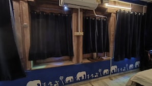 Une pièce aux murs en bois, une fenêtre avec des rideaux, un climatiseur fixé au mur et un mur bleu orné de silhouettes d’éléphants.