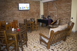 A room with a brick wall, a television mounted on it, wooden furniture including a table and chairs, and a sofa. - Hostel Cafayate (Cafayate)
