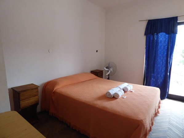 Quarto com uma cama, mesa de cabeceira de madeira, ventilador fixado na parede e janela com cortinas azuis.