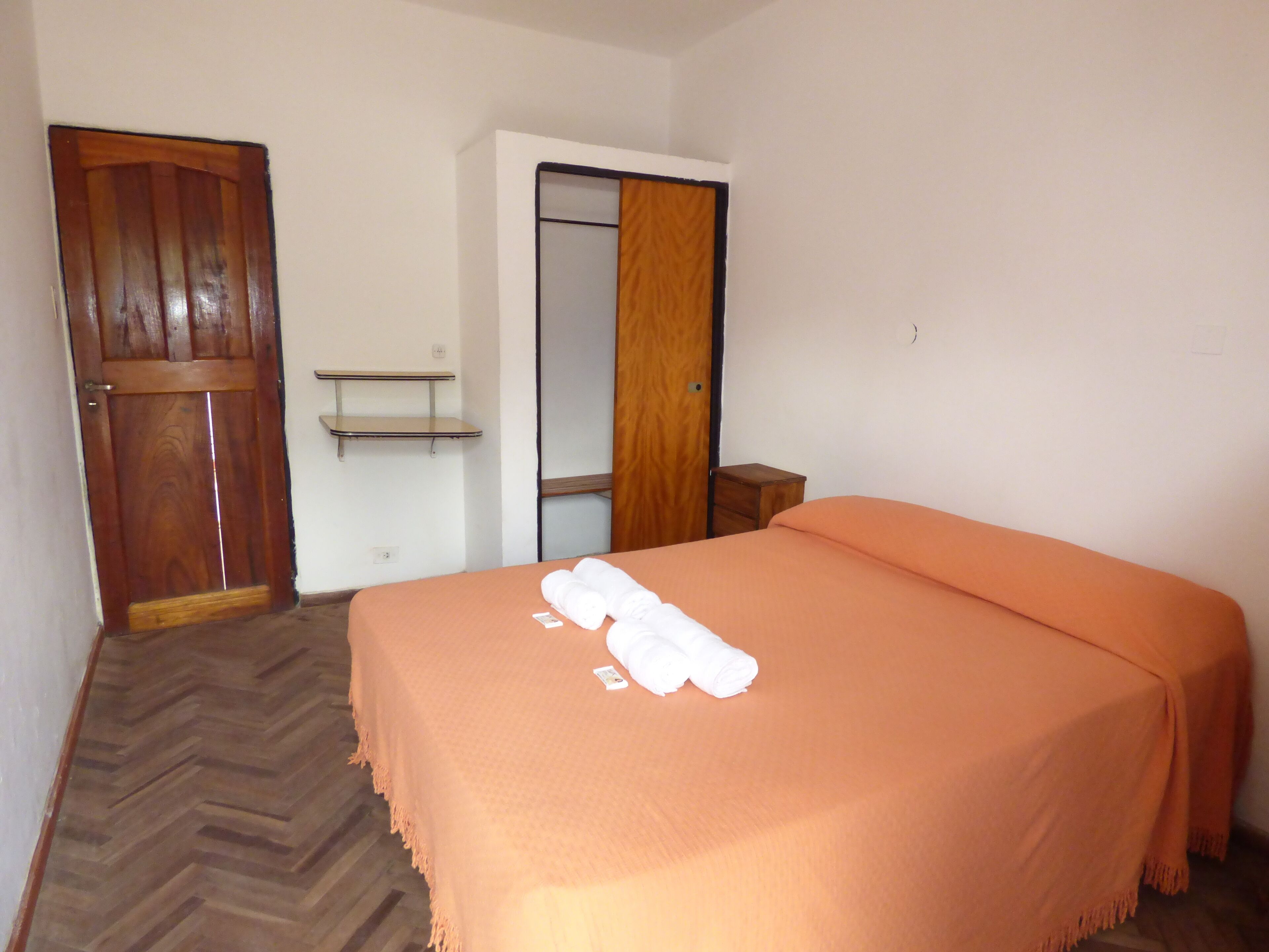 A hotel room with a wooden door, a wardrobe, a bed with an orange bedspread, and a small wooden nightstand.