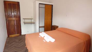 A hotel room with a wooden door, a wardrobe, a bed with an orange bedspread, and a small wooden nightstand.