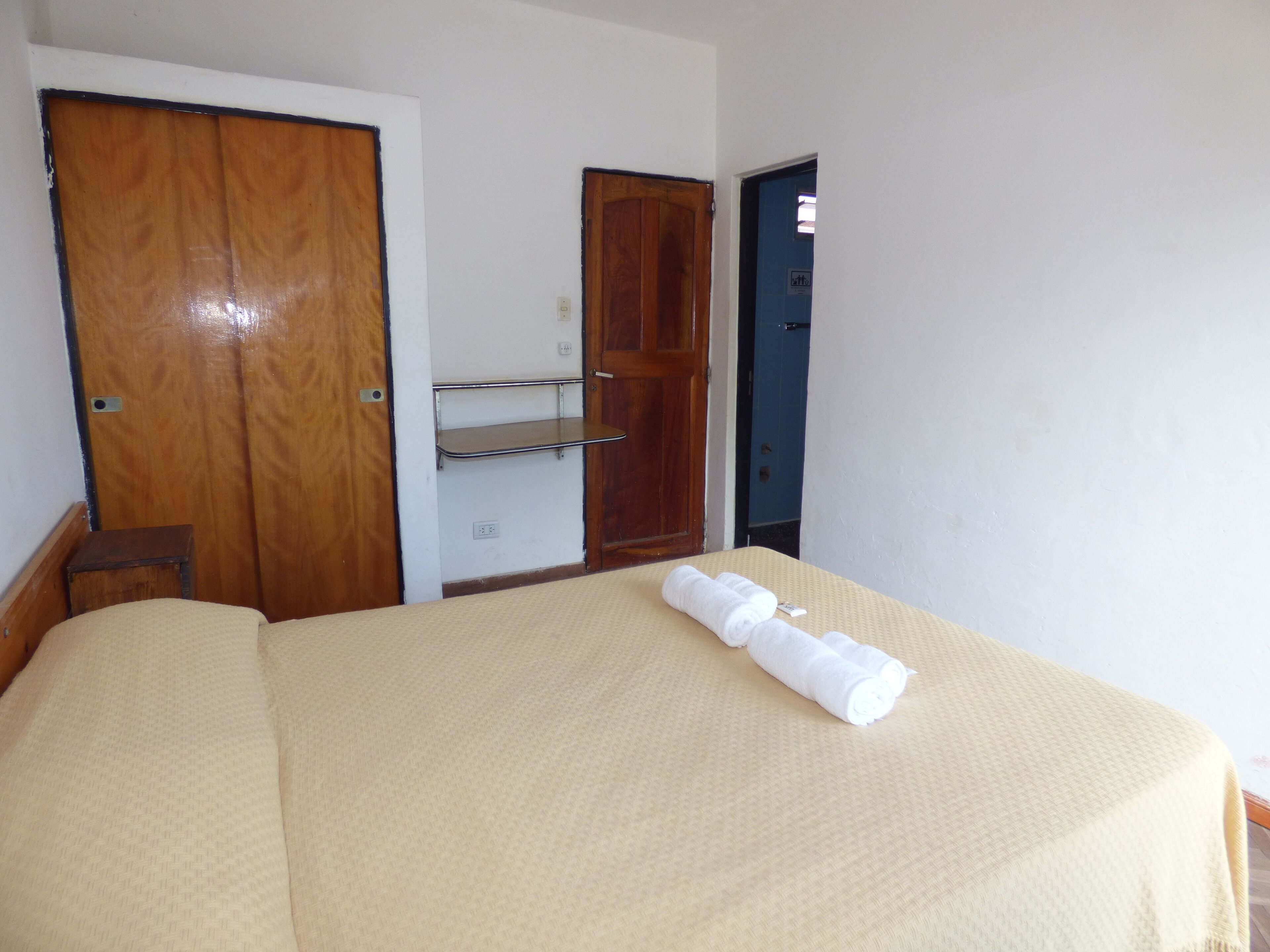 A hotel room with a bed, a wooden wardrobe, a shelf, and a door.