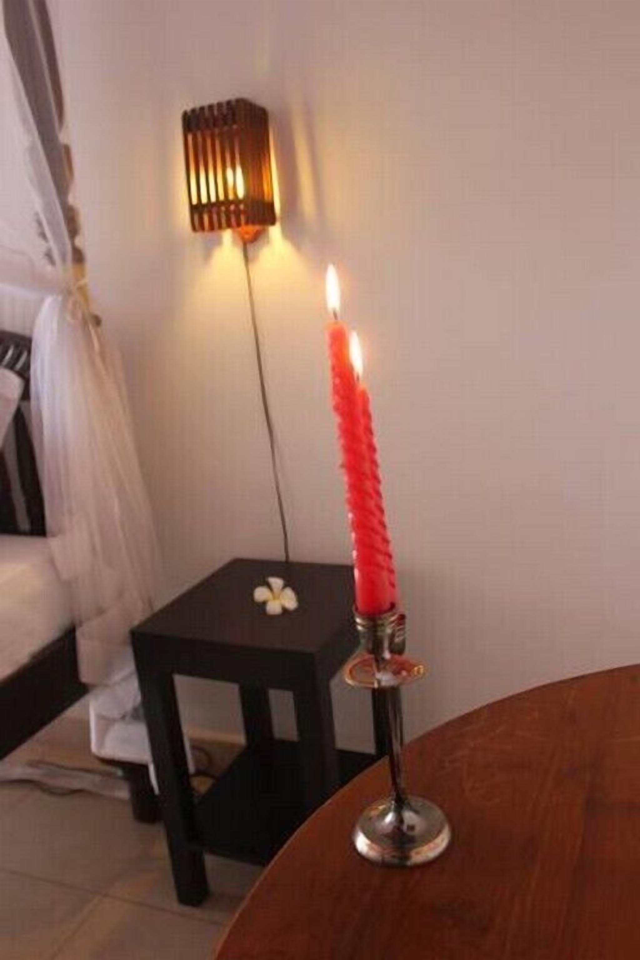 Une chambre avec un lit, une table de chevet sur laquelle se trouve une bougie rouge, et un luminaire fixé au mur.