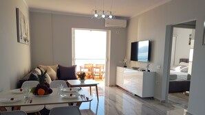A modern living room with a dining area, a flat-screen TV mounted on the wall, and a balcony with wooden furniture. - Apartment in front of the beach (Sarandë)