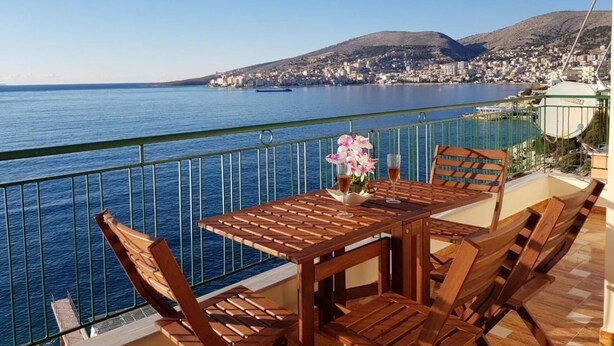 A wooden table with chairs on a balcony overlooking a body of water with a city and mountains in the background. - Apartment in front of the beach (Sarandë)
