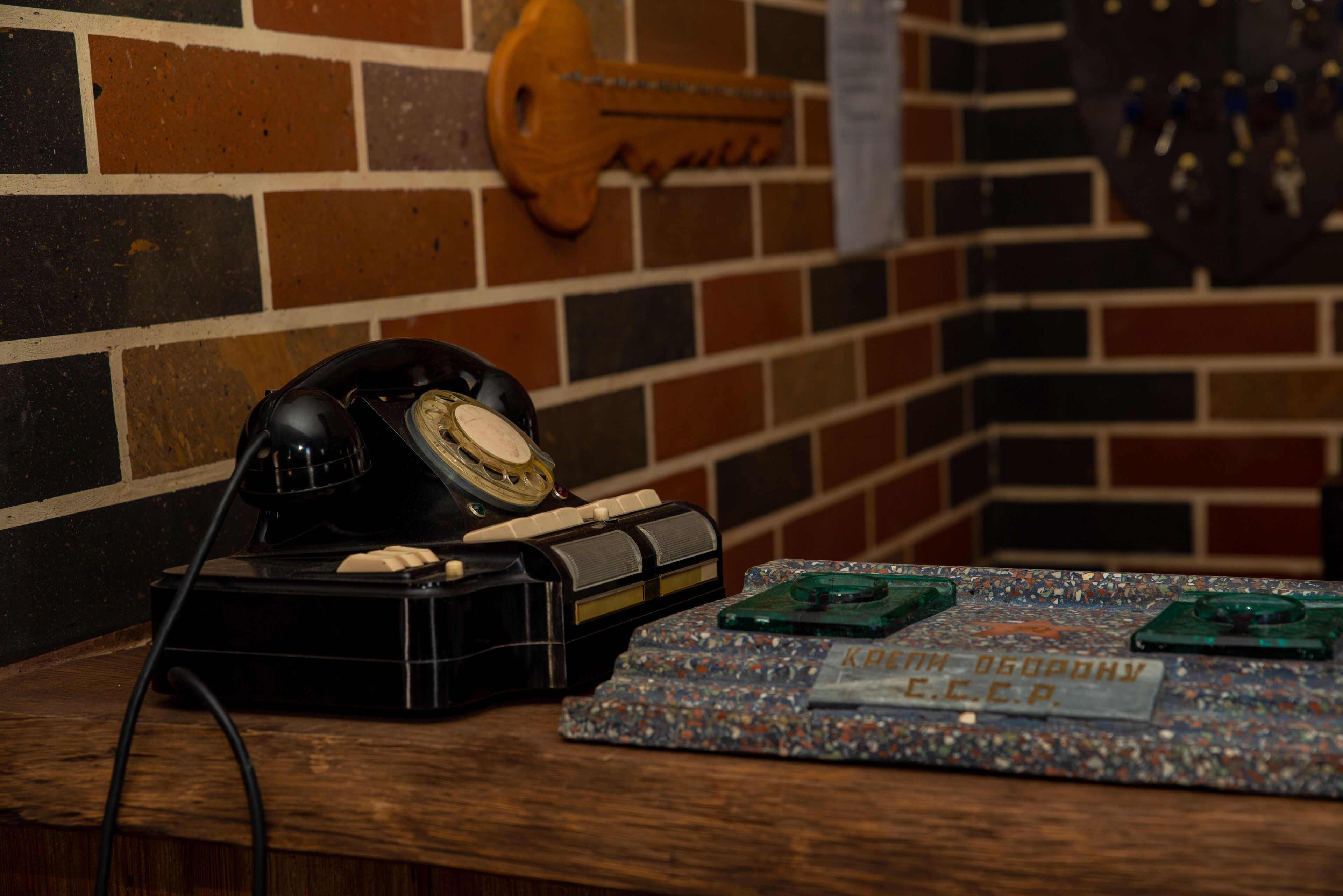 Un vieux téléphone à cadran rotatif posé sur une table en bois, une clé décorative et une plaque nominative avec du texte en cyrillique.
