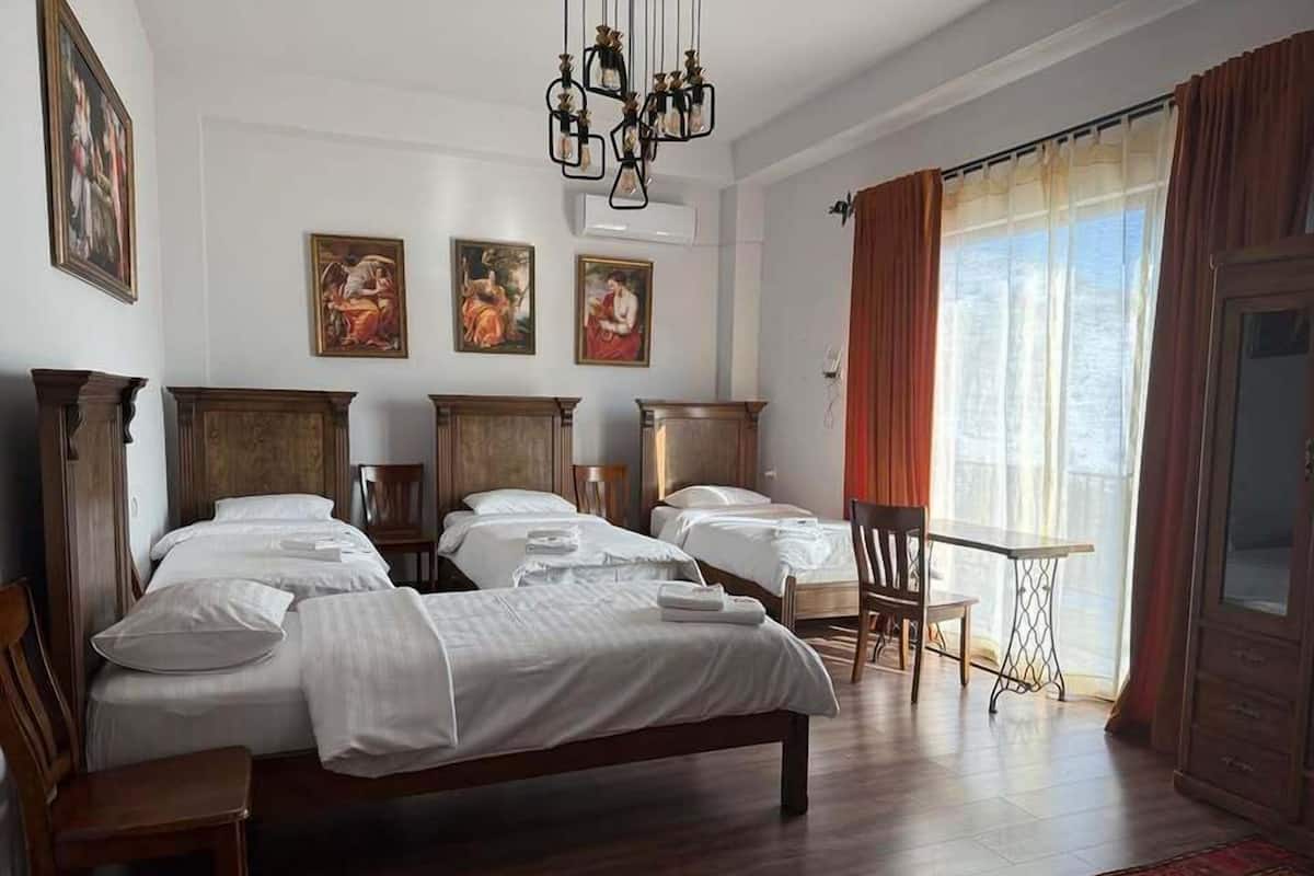 Habitación de hotel con una cama grande, dos camas más pequeñas, un escritorio y una silla. Hay tres cuadros en la pared, una araña y una ventana con cortinas.