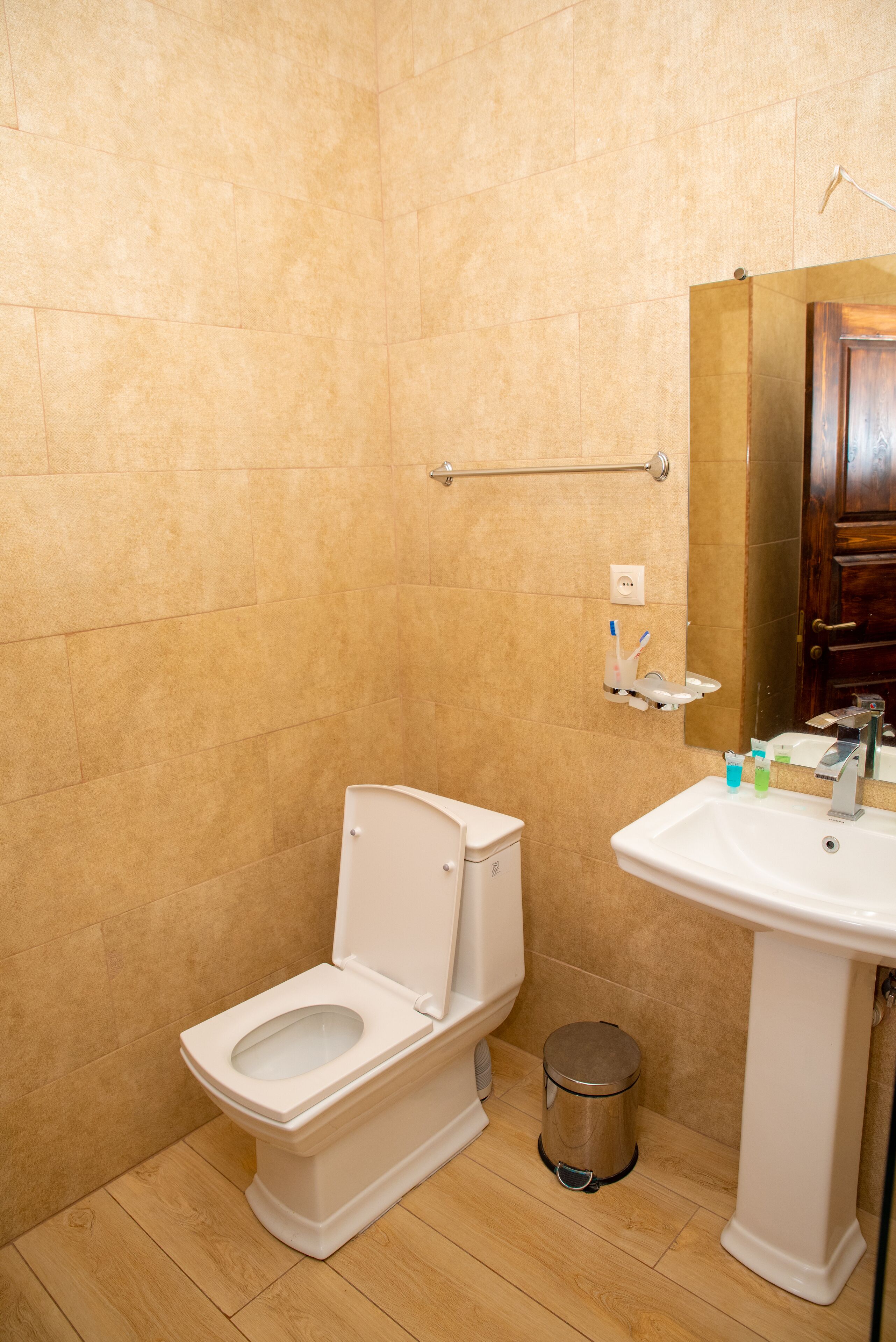 Une salle de bain avec des carreaux de couleur beige, des toilettes blanches, un lavabo sur colonne et une petite poubelle.
