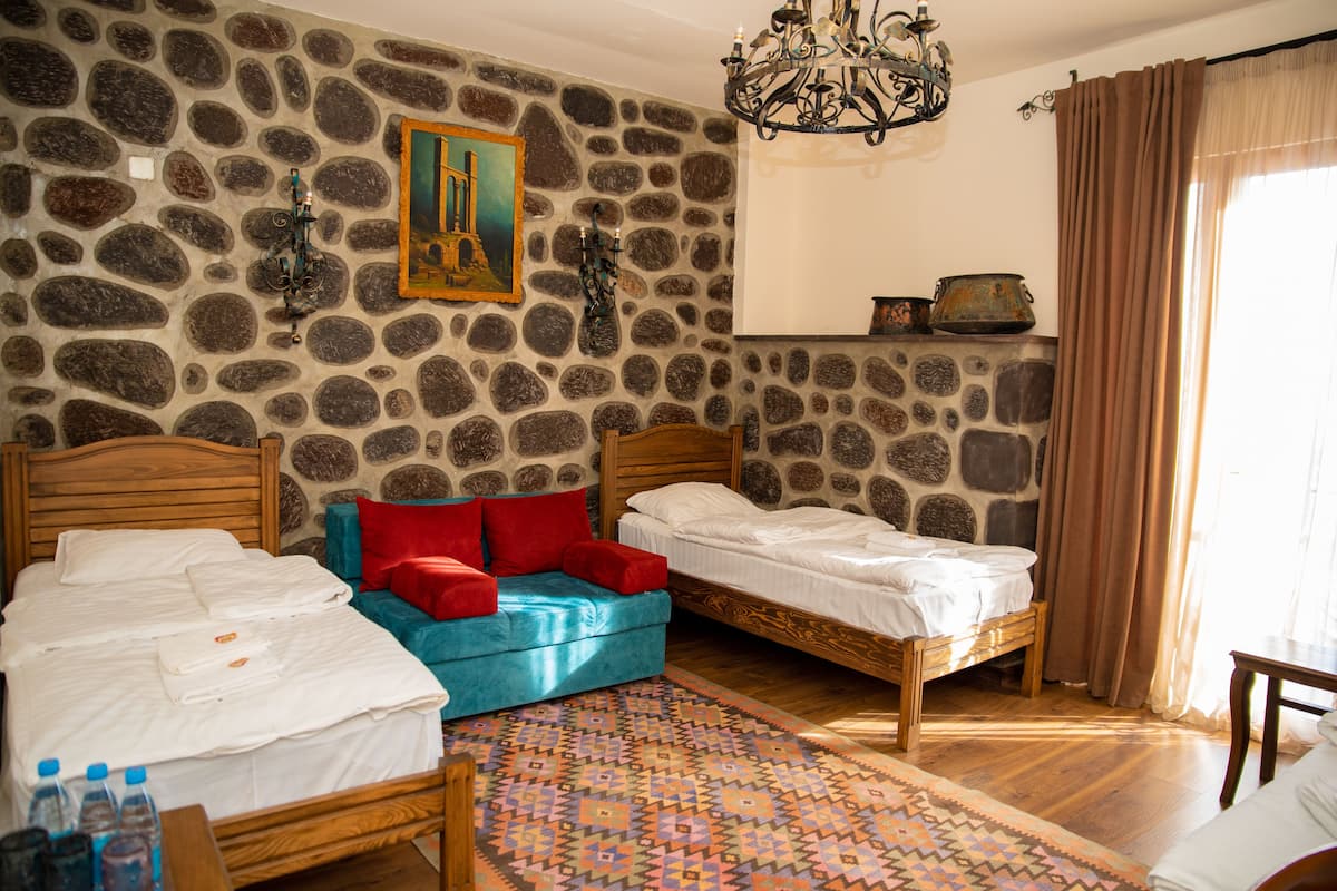 Una habitación con dos camas de madera, un sofá azul, un tapete estampado, una pared de piedra, una araña y un cuadro.