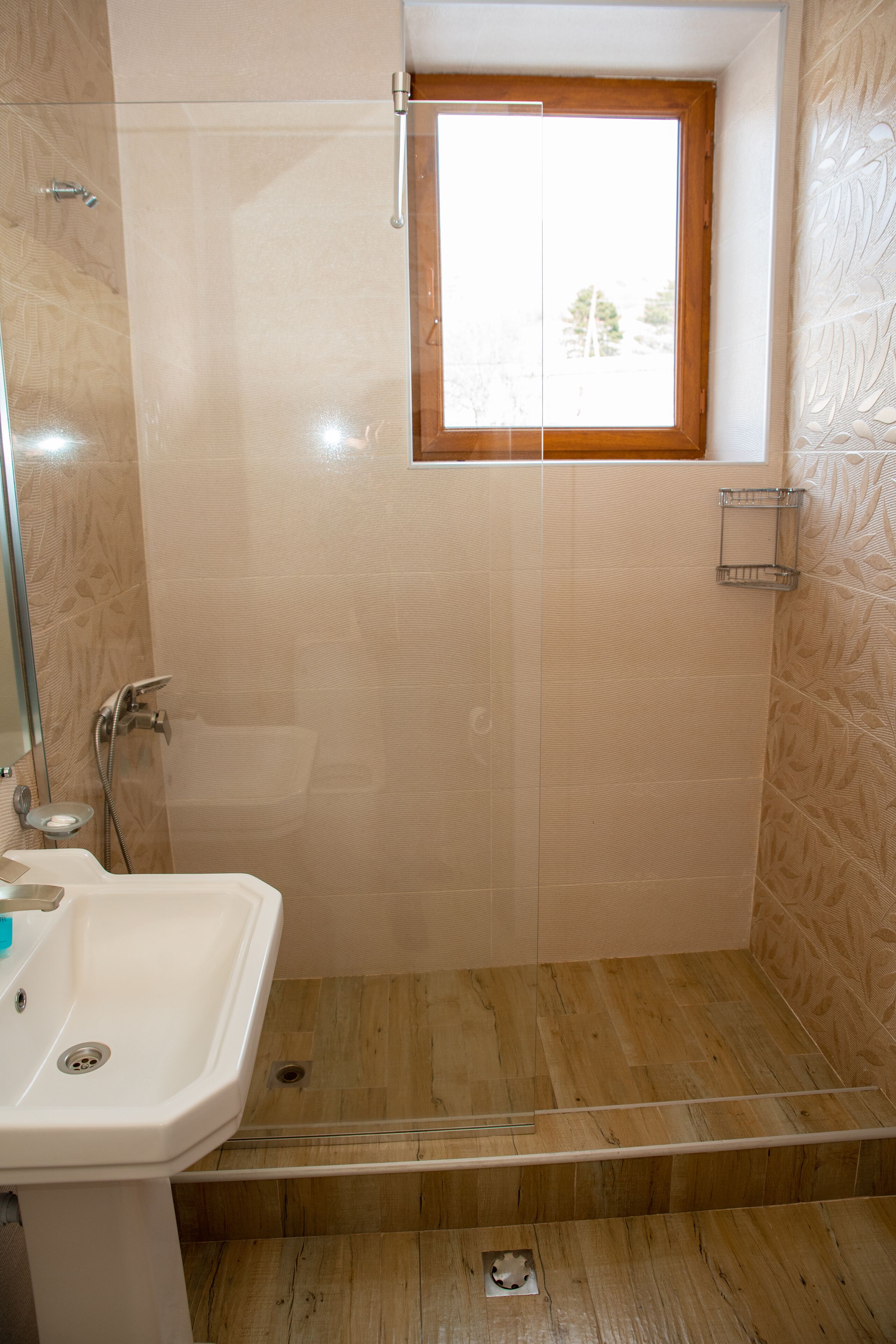 Un baño con cabina de ducha de vidrio, suelo de madera, lavamanos blanco y una ventana pequeña.