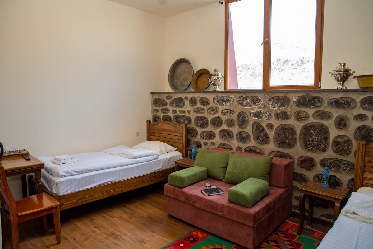 Un dormitorio con una cama individual, una mesita de noche de madera, un sofá rojo con cojines verdes, una pared de piedra y una ventana con vistas a la nieve.