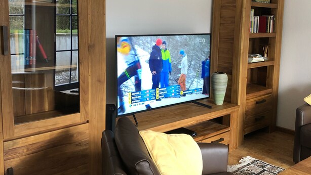 A living room with a television displaying a sports event, wooden furniture, and a brown leather sofa. - Maison Euba am Zeisigwald (Euba)