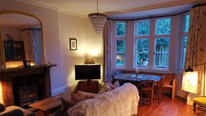 Ein gemütliches Wohnzimmer mit Kamin, Fernseher, Esstisch und einem großen Fenster mit Blick auf Grünflächen.