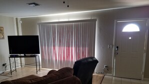 Una sala de estar con una televisión de pantalla plana sobre un mueble bajo, un sofá marrón, una silla de oficina negra y una puerta con ventanal.