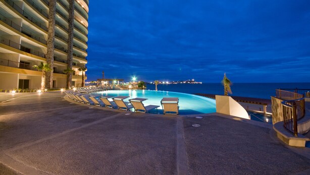Zona de piscina de hotel con sillones de descanso y vista al océano al atardecer.